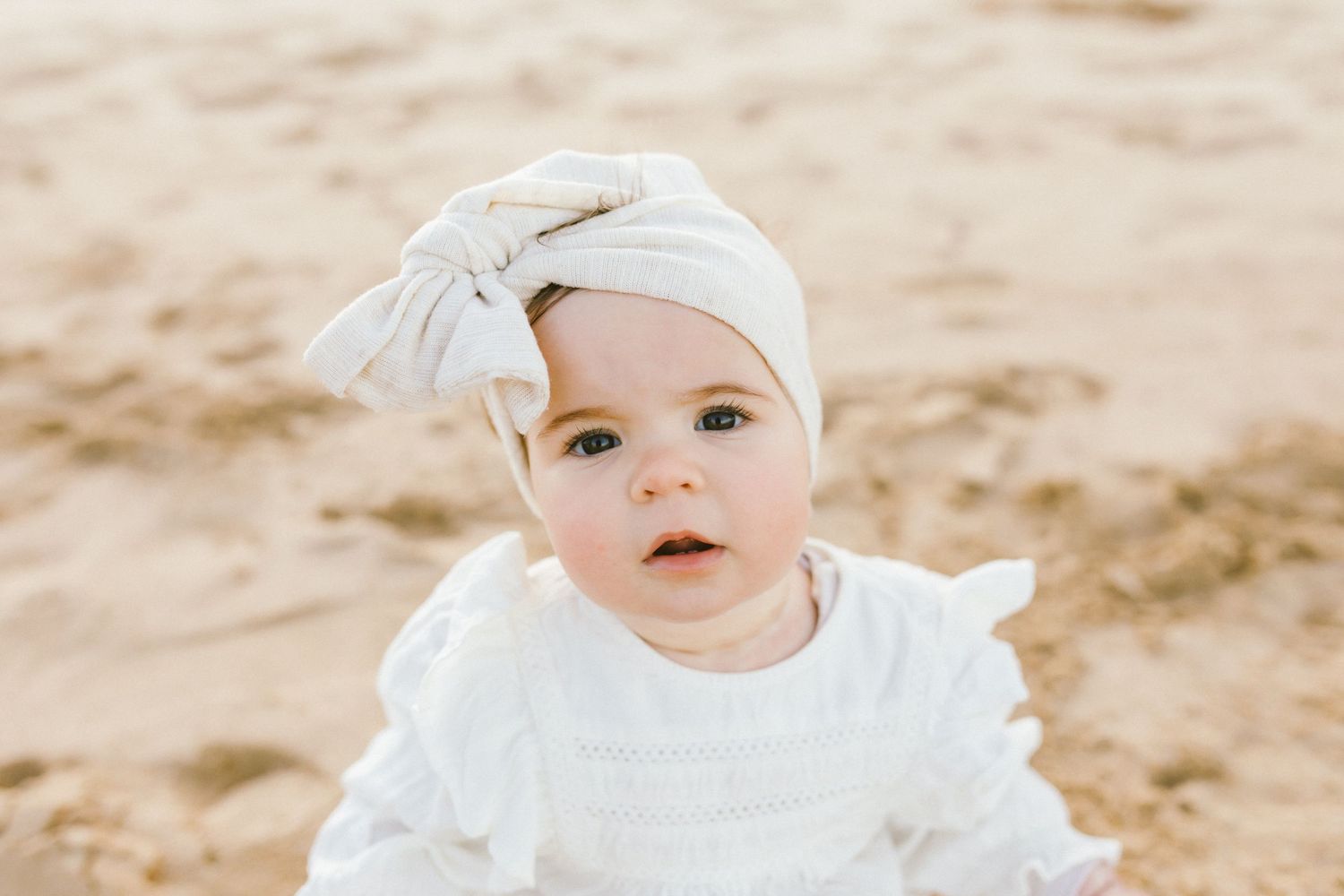 A small baby wearing white clothing and a white hat sits on a sandy beach in soft natural light.