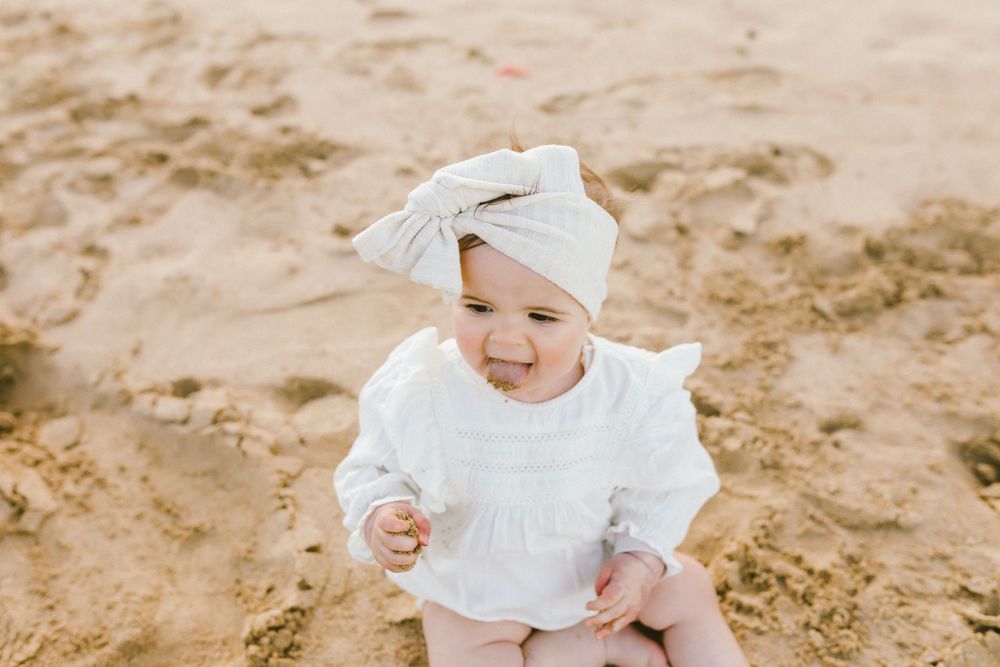 A small child in a white dress and bow sits on sandy ground playing in a collage of beach photos.