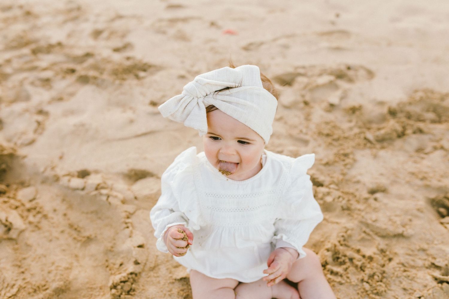 A small child in a white dress and bow sits on sandy ground playing in a collage of beach photos.