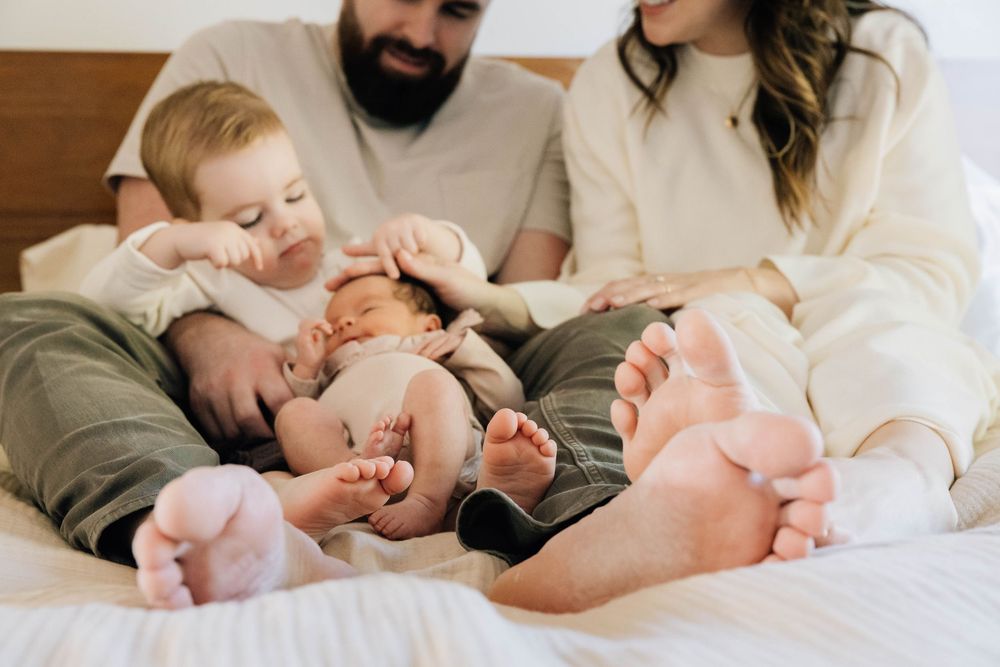 Family with toddler and newborn relaxing together on a white bed, showing tiny baby feet.