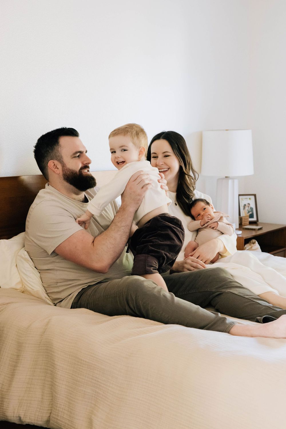 Happy family of four sitting on a bed, parents smiling as toddler laughs and mom holds newborn.