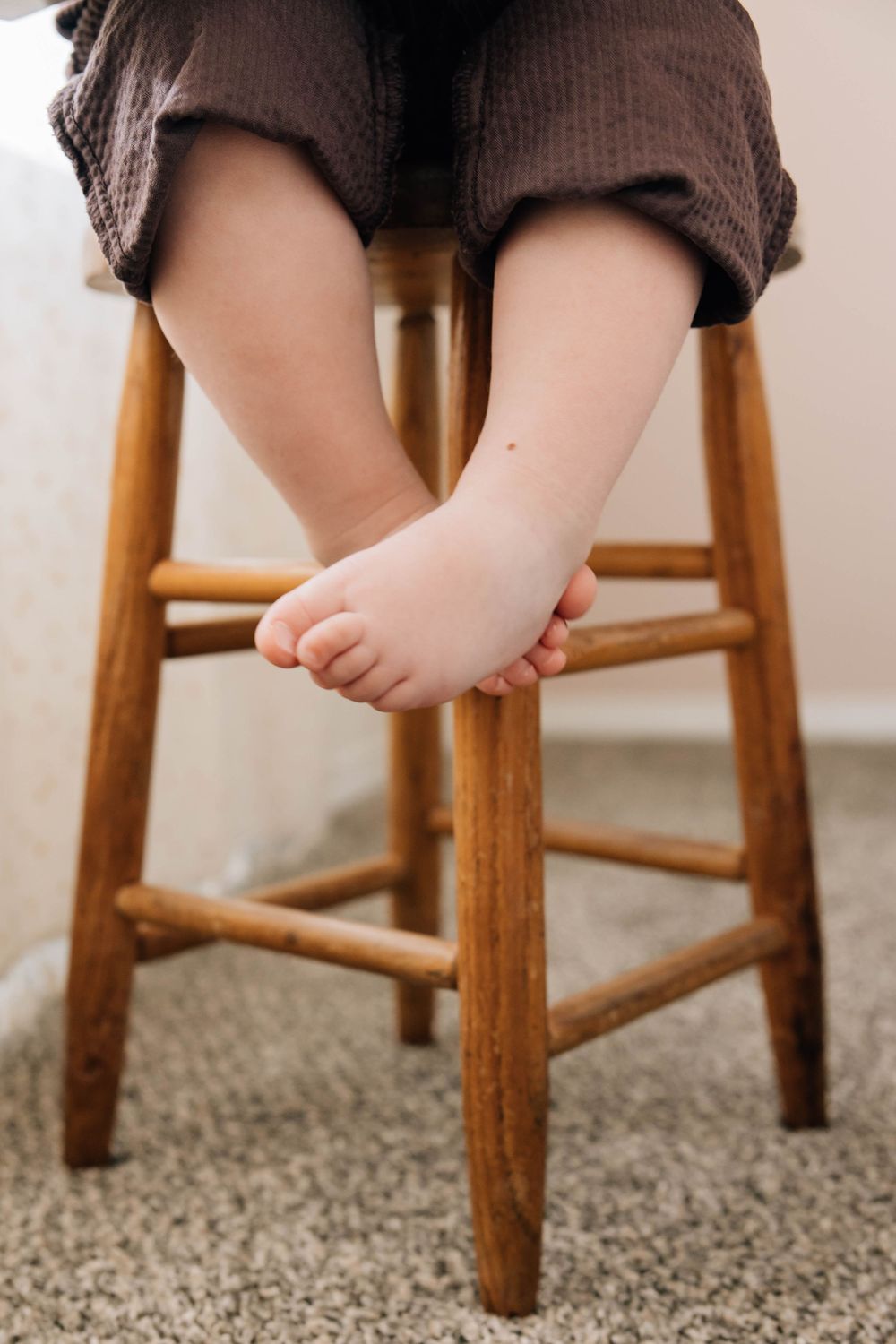 Close-up of a toddler's bare feet dangling from a wooden stool on a carpeted floor.