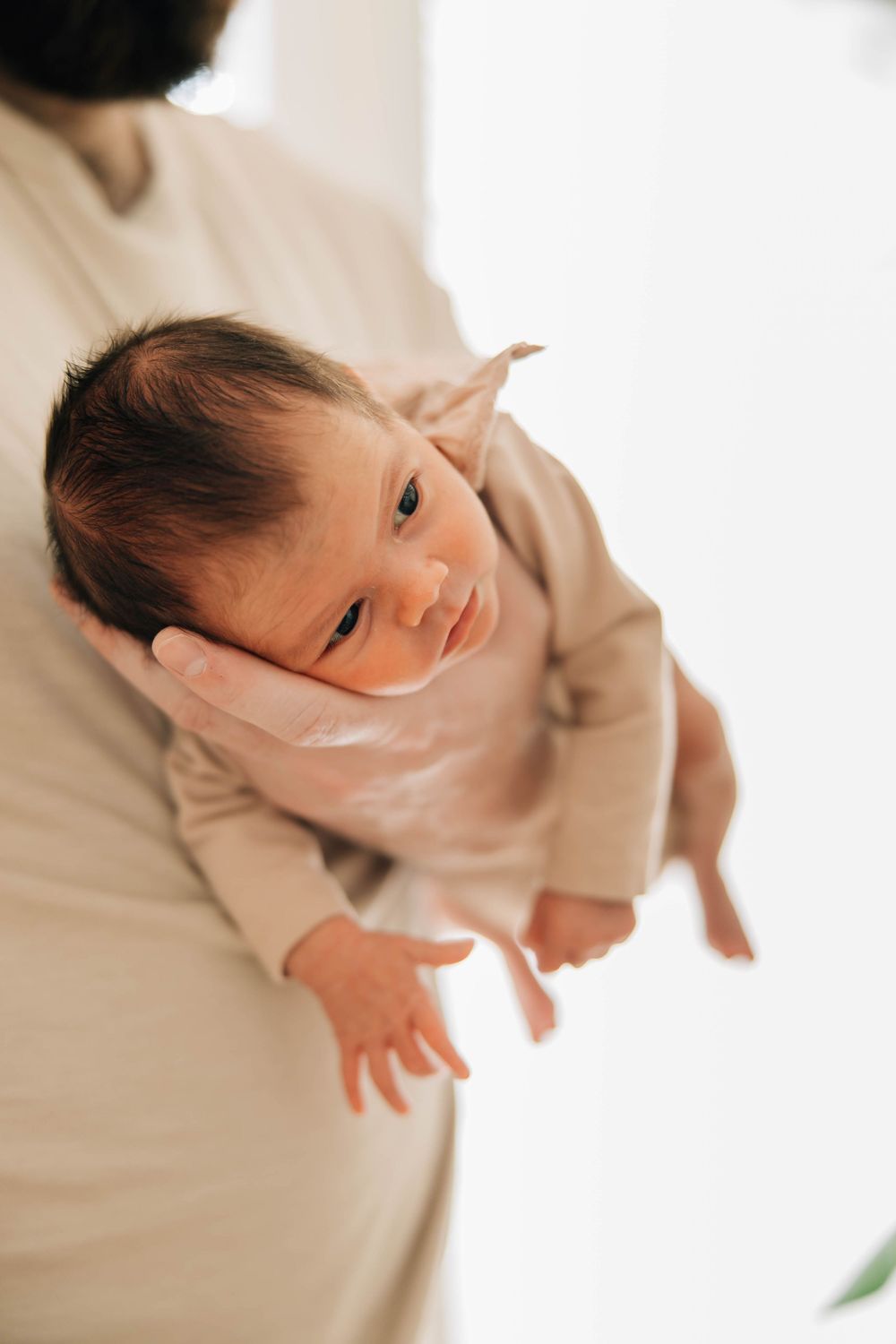 A newborn baby in a beige onesie being cradled in a parent's arms against a bright background.