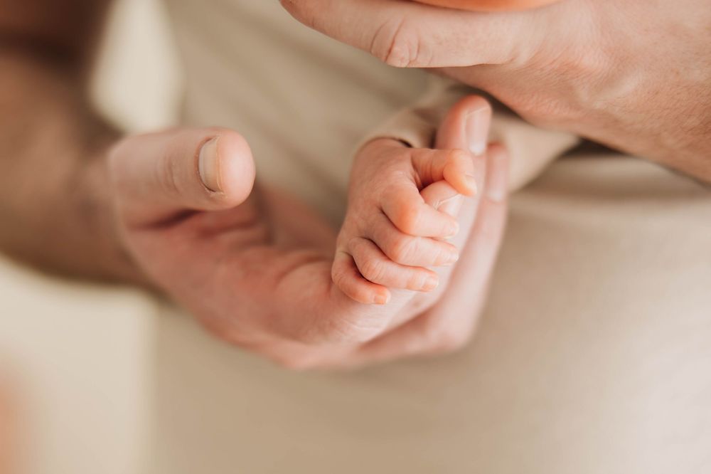 A parent's hands gently holding a newborn baby's tiny fist in a tender close-up photo.