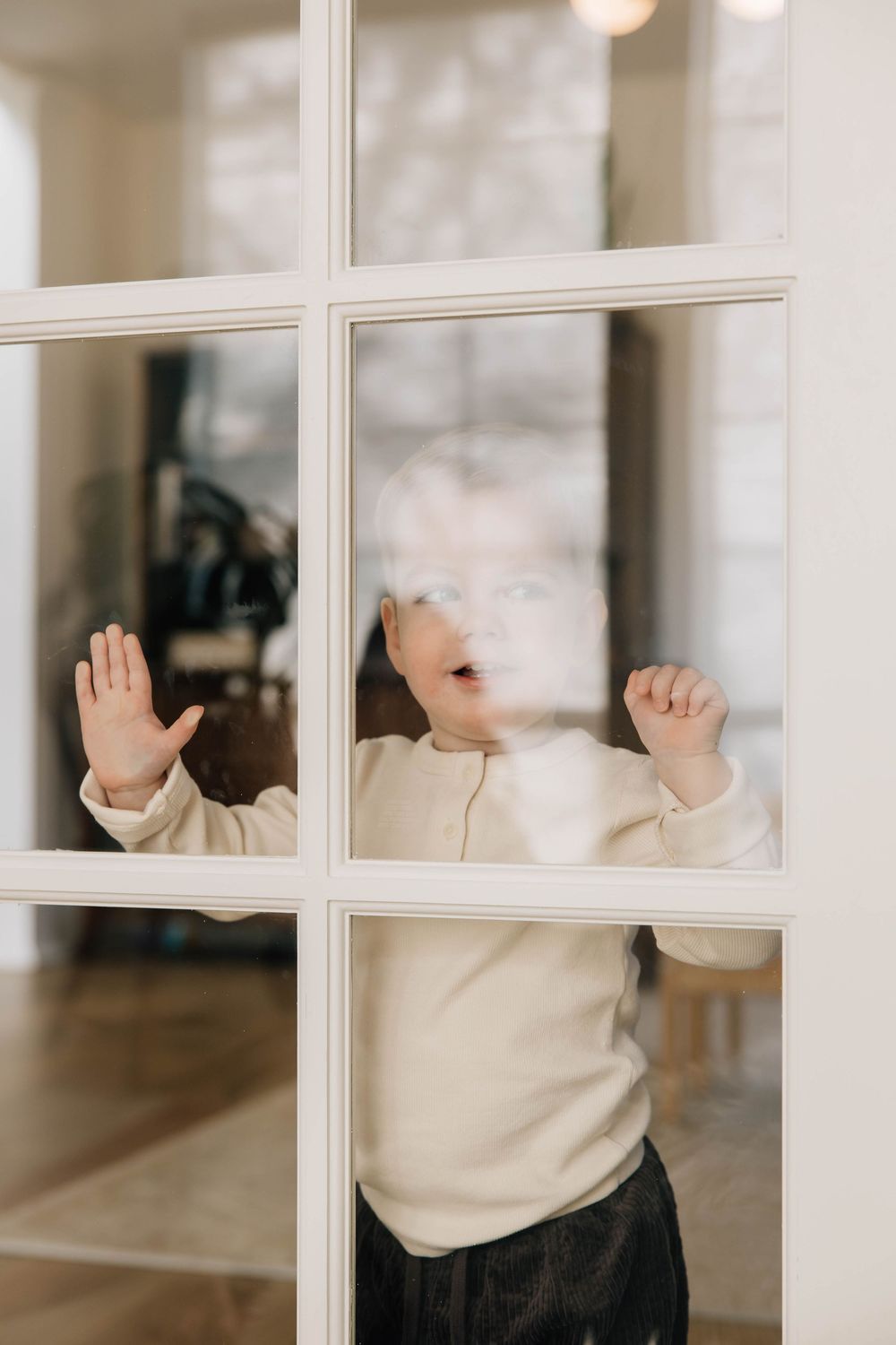 A toddler in a white sweater pressing hands and face against a white-framed glass door.