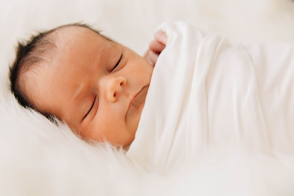 Peaceful newborn baby sleeping soundly wrapped in a white swaddle on a fluffy white blanket.