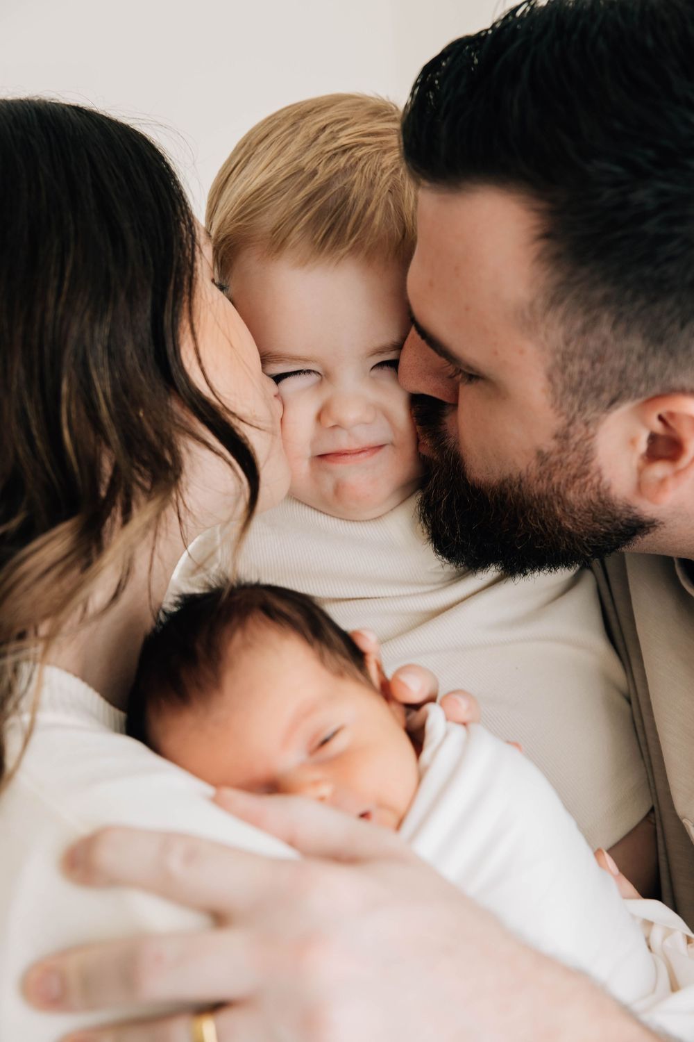 Happy family with bearded father and mother kissing toddler while holding sleeping newborn baby.