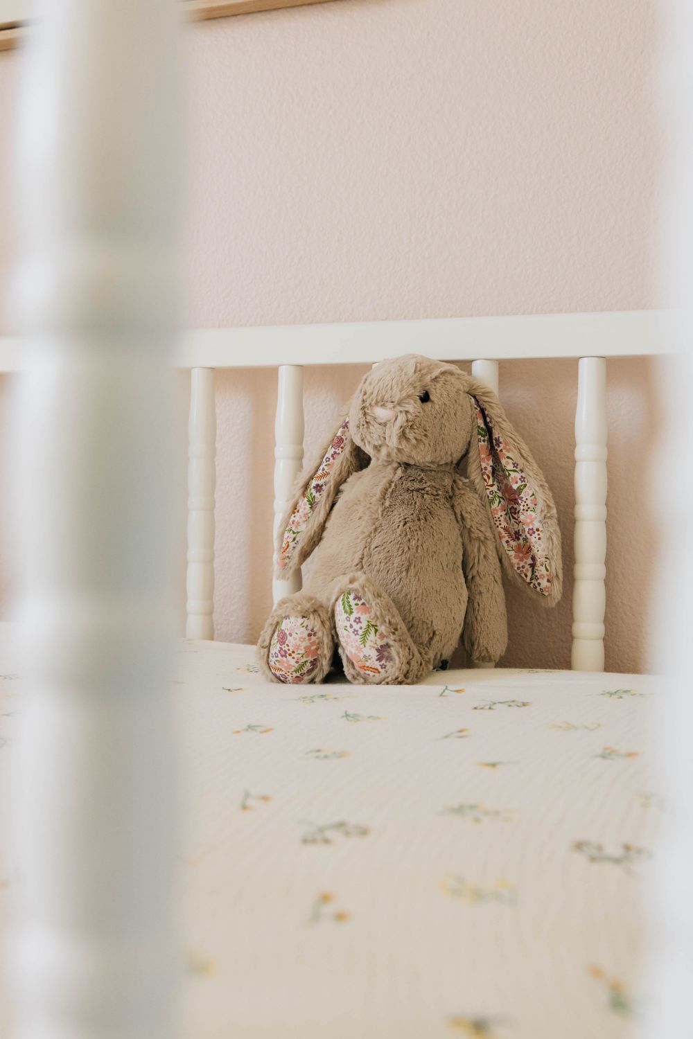Jellycat bunny stuffed animal sitting in white wooden baby crib with floral sheet.