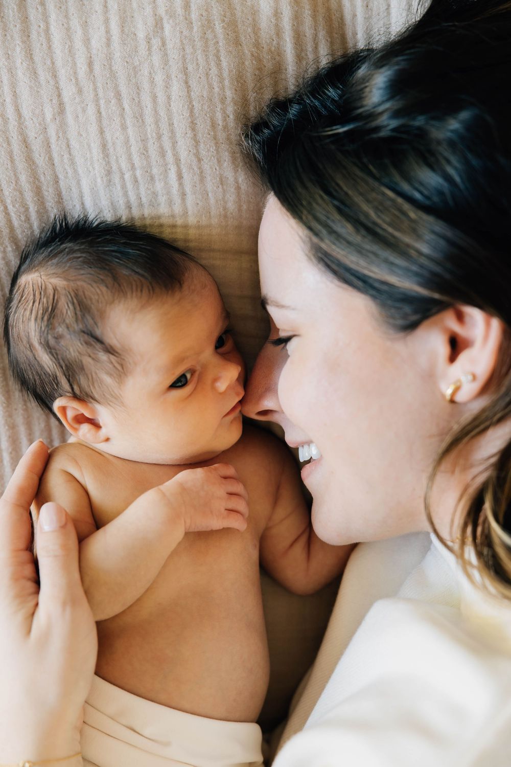 Smiling dark-haired mother lying face-to-face with alert newborn baby on beige blanket.