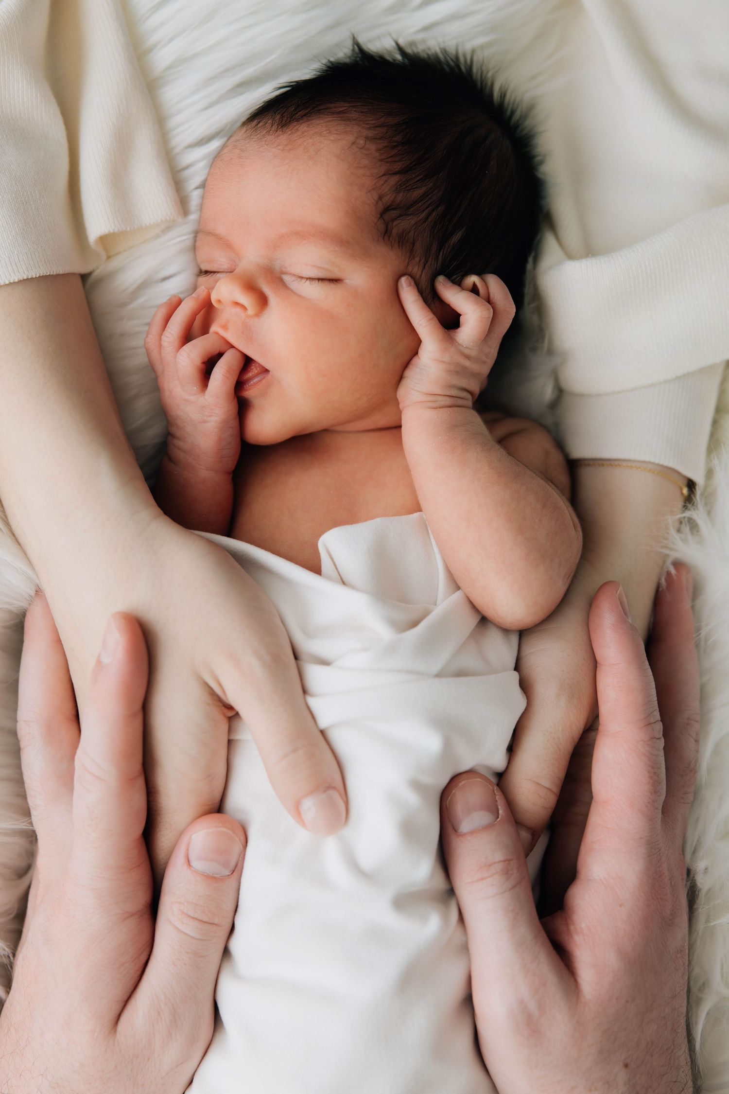 Sleeping newborn baby in white wrap cradled gently by parents hands from above.