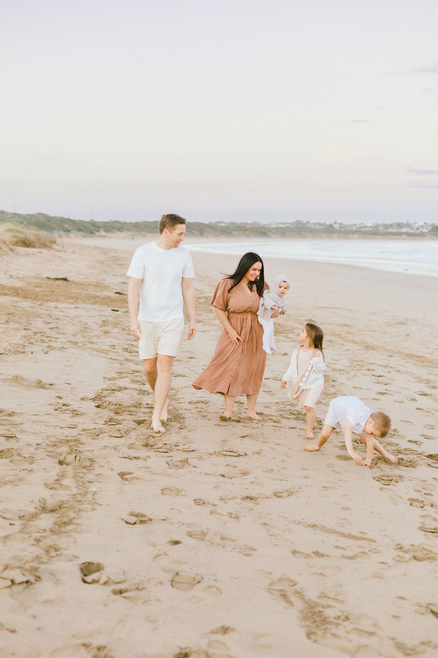 A family walks together along a sandy beach at sunset in warm neutral tones.