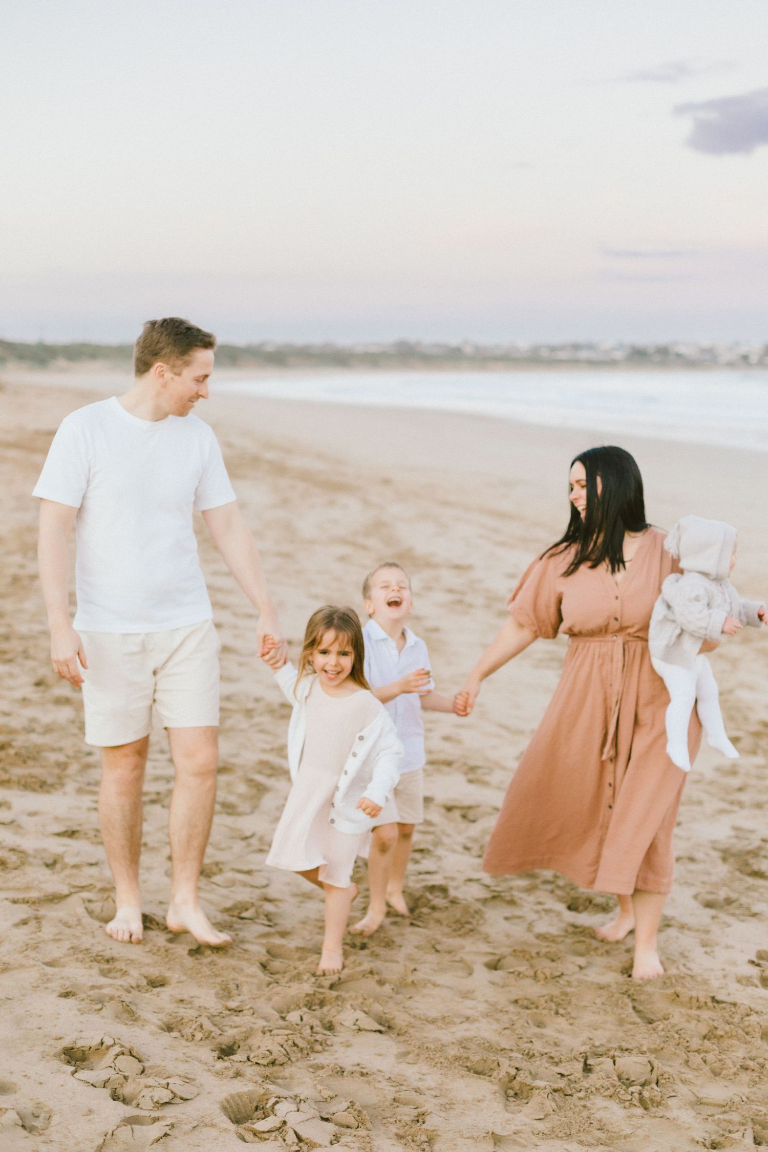 A family walks together holding hands along a sandy beach at sunset in soft pastel tones.
