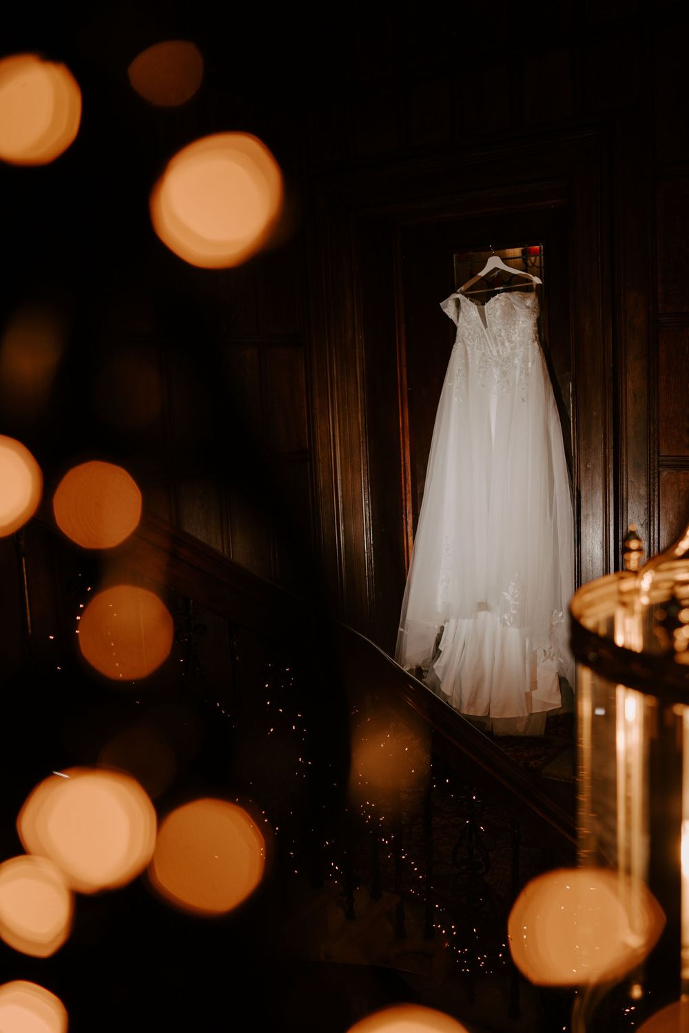 Elegant white wedding dress hangs in a dark room with warm bokeh lighting effects creating a romantic ambiance.