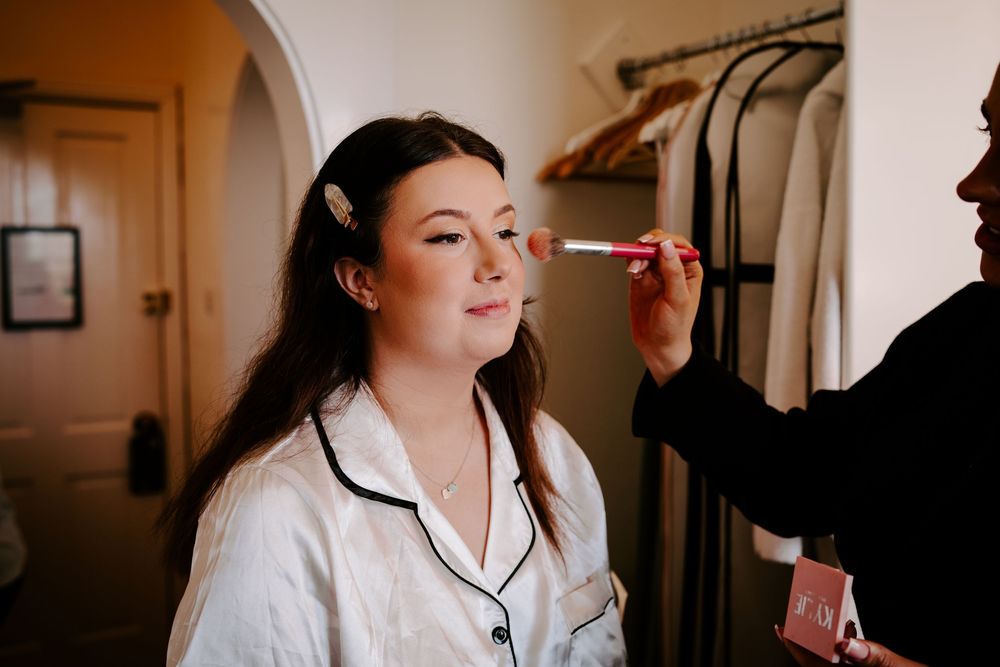 A makeup artist applies cosmetics to a person wearing a white satin robe with black piping in a warmly lit room.
