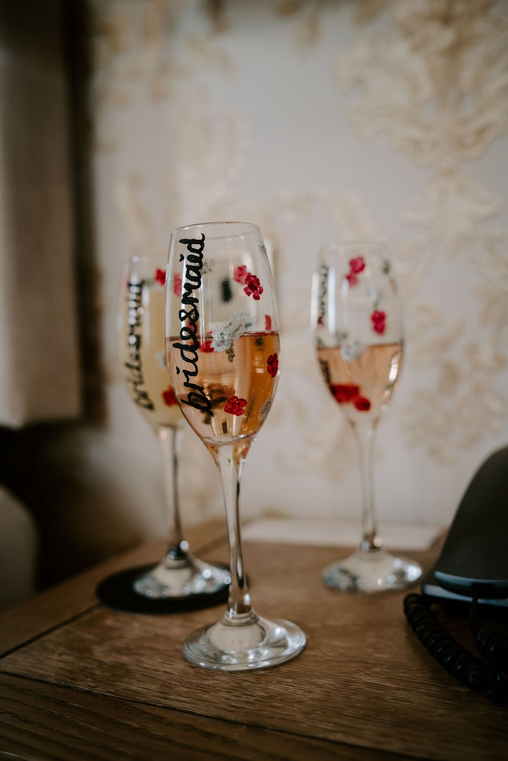 Champagne flutes with bridesmaid text and decorative red accents sitting on wooden surface.