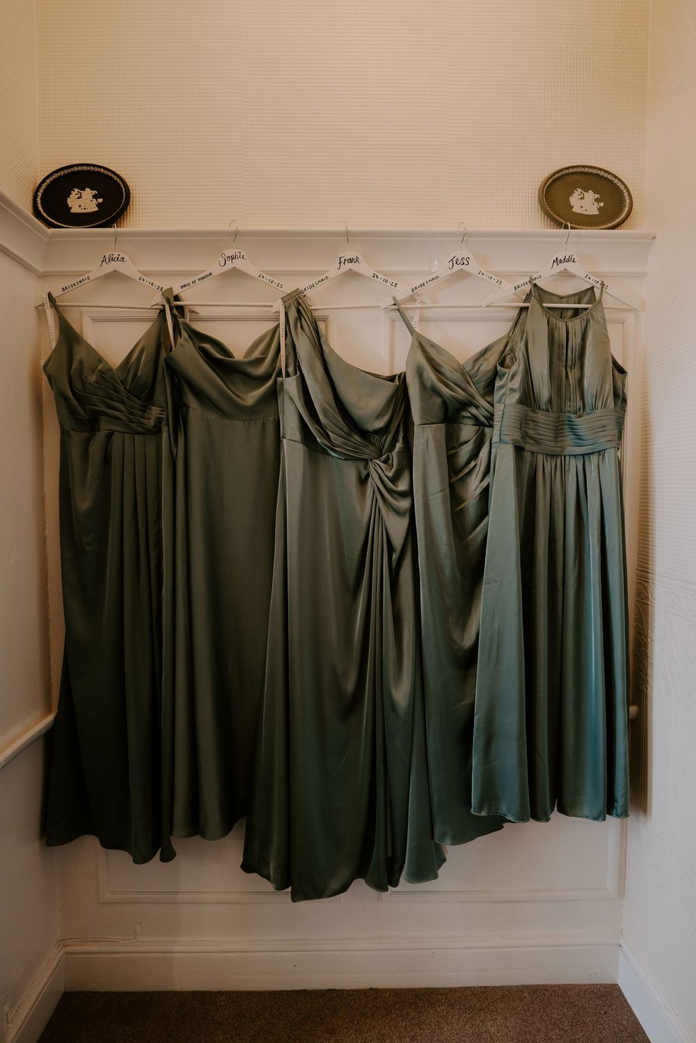 Sage green bridesmaid dresses hanging in a row on a white wall.