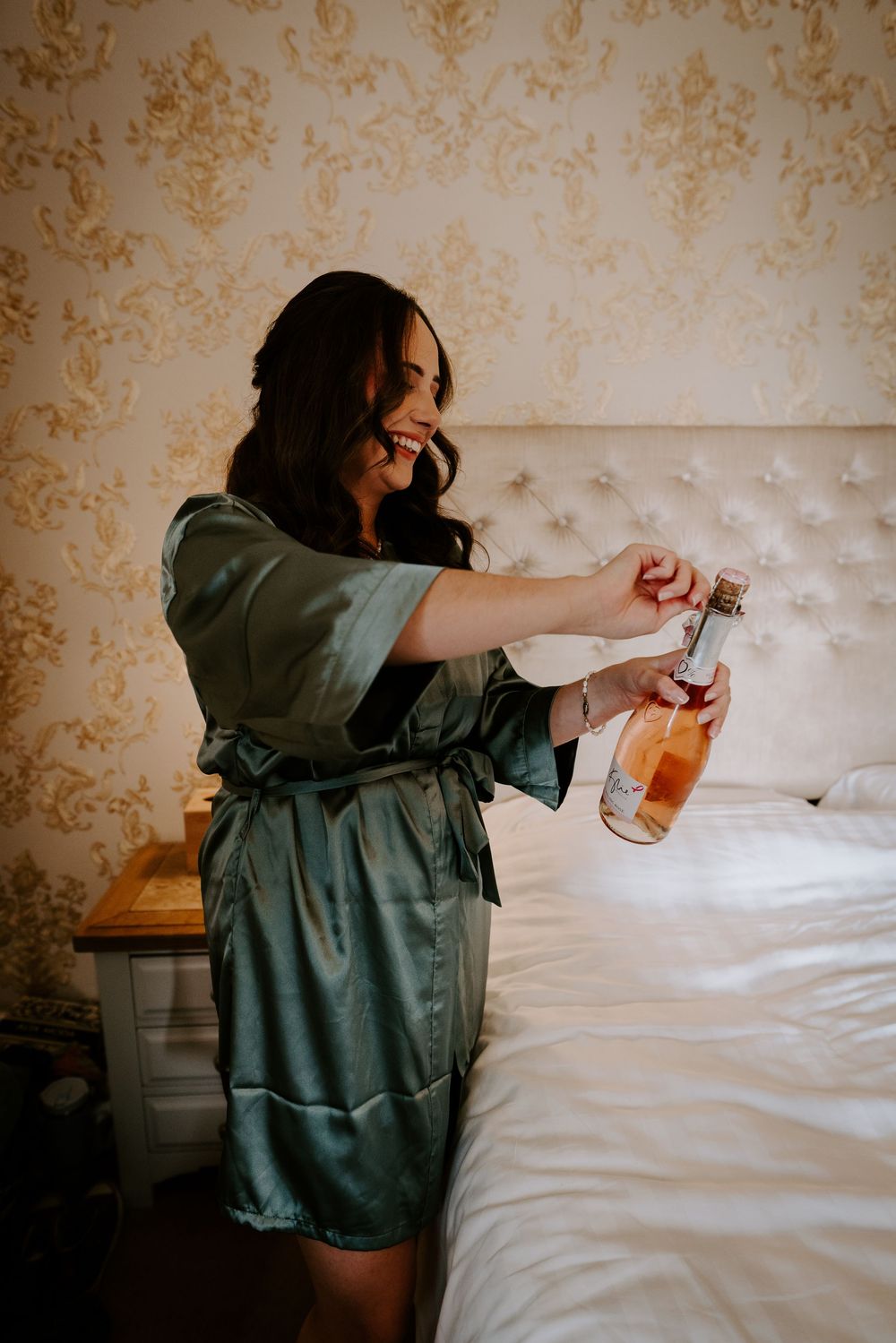 Someone wearing a sage green silk robe holds champagne glasses on a bed with floral wallpaper.