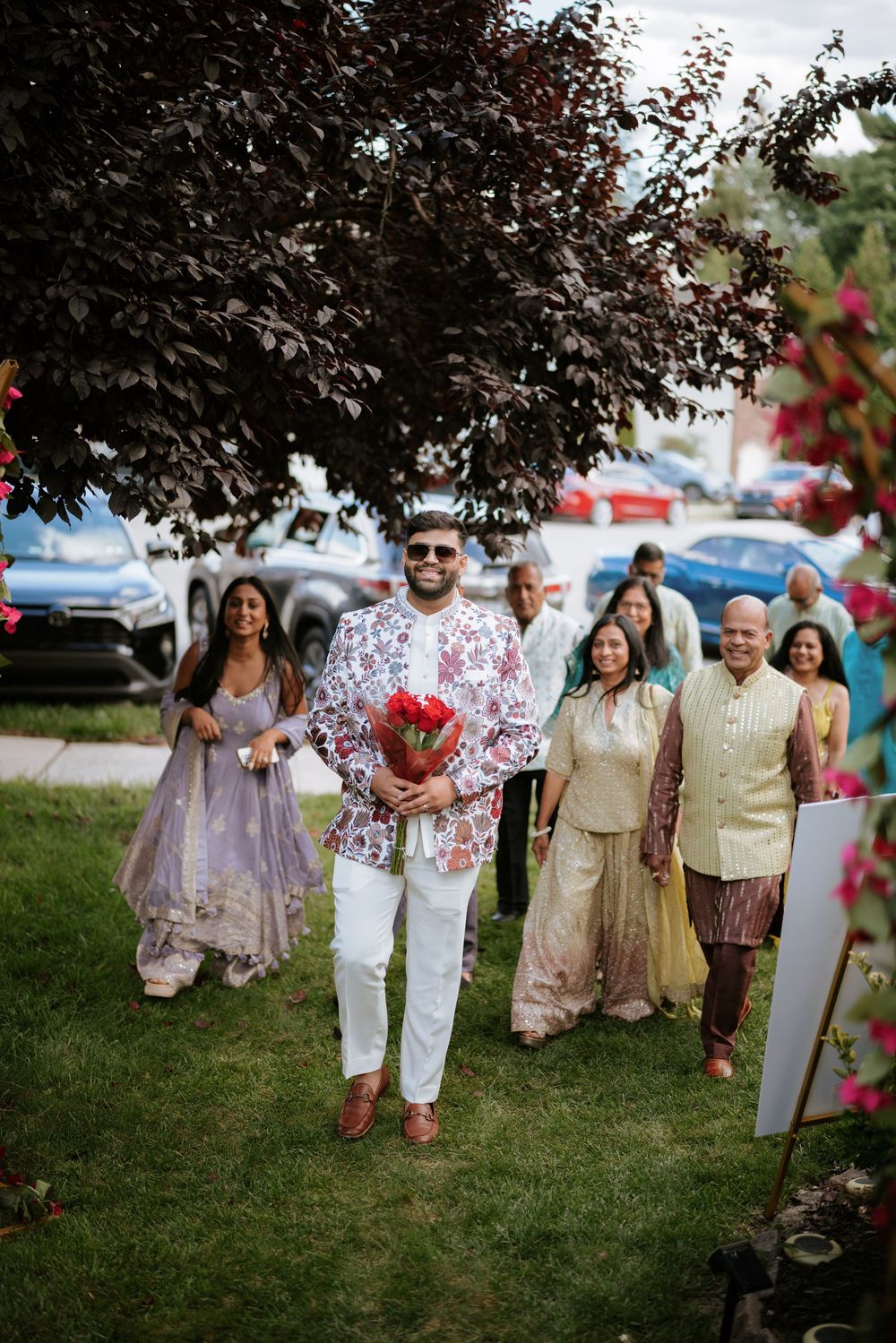 Wedding guests in traditional Indian and fusion attire pose together on a grassy lawn during an outdoor celebration.