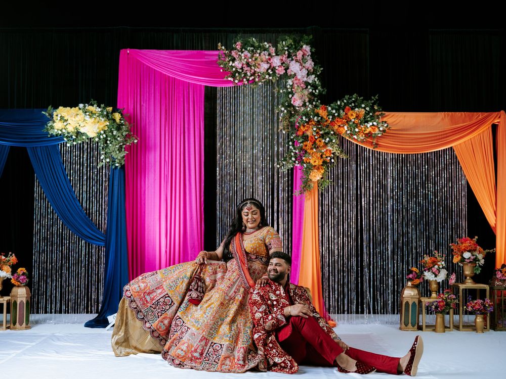 Elegant Indian wedding stage decor featuring bright pink and orange drapes with floral arrangements against a sparkly backdrop.