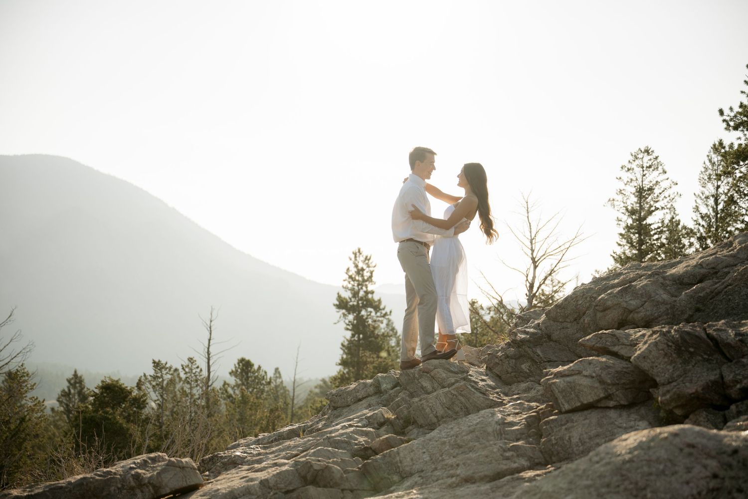 Two people share a moment while standing on a rocky mountain overlook with misty peaks in the background.