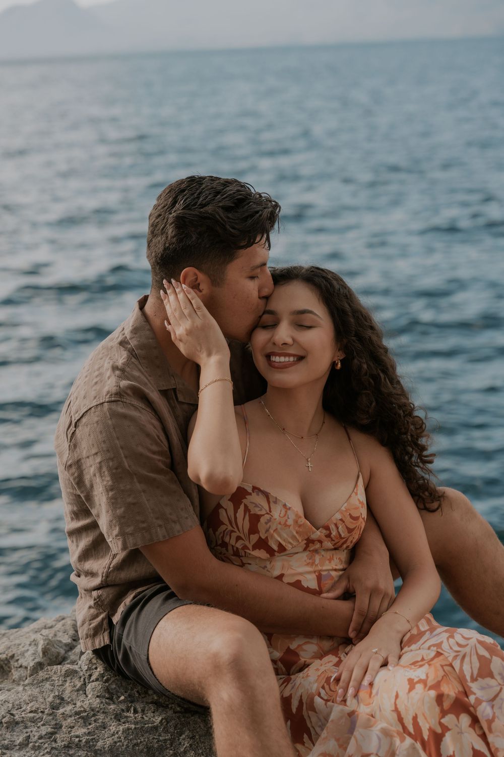 A couple sits on rocks by the ocean, sharing a romantic kiss on the cheek while the woman smiles blissfully.