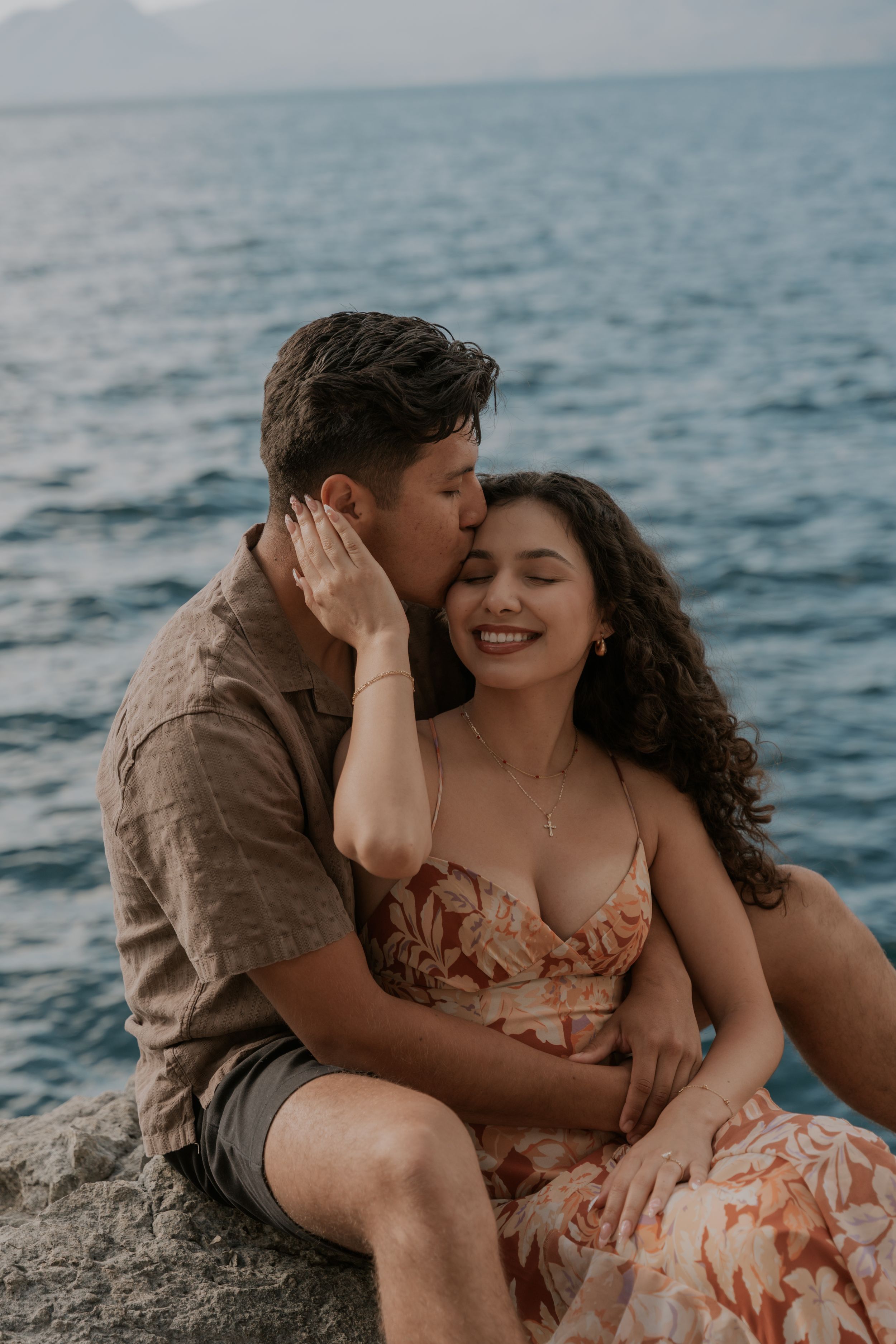 A couple sits on rocks by the ocean, sharing a romantic kiss on the cheek while the woman smiles blissfully.