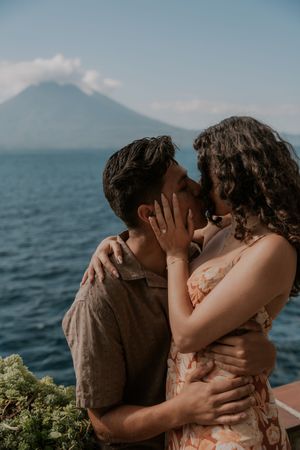 Couple sharing a romantic kiss with a volcanic mountain and blue lake in the scenic background.