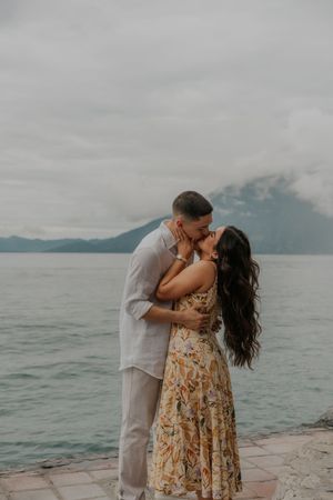 A couple embraces and kisses on a stone pier overlooking a serene lake with mountains in the background.