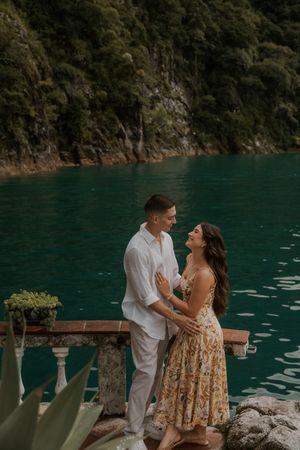 A romantic couple shares a tender moment on a wooden deck overlooking turquoise waters surrounded by lush mountains.