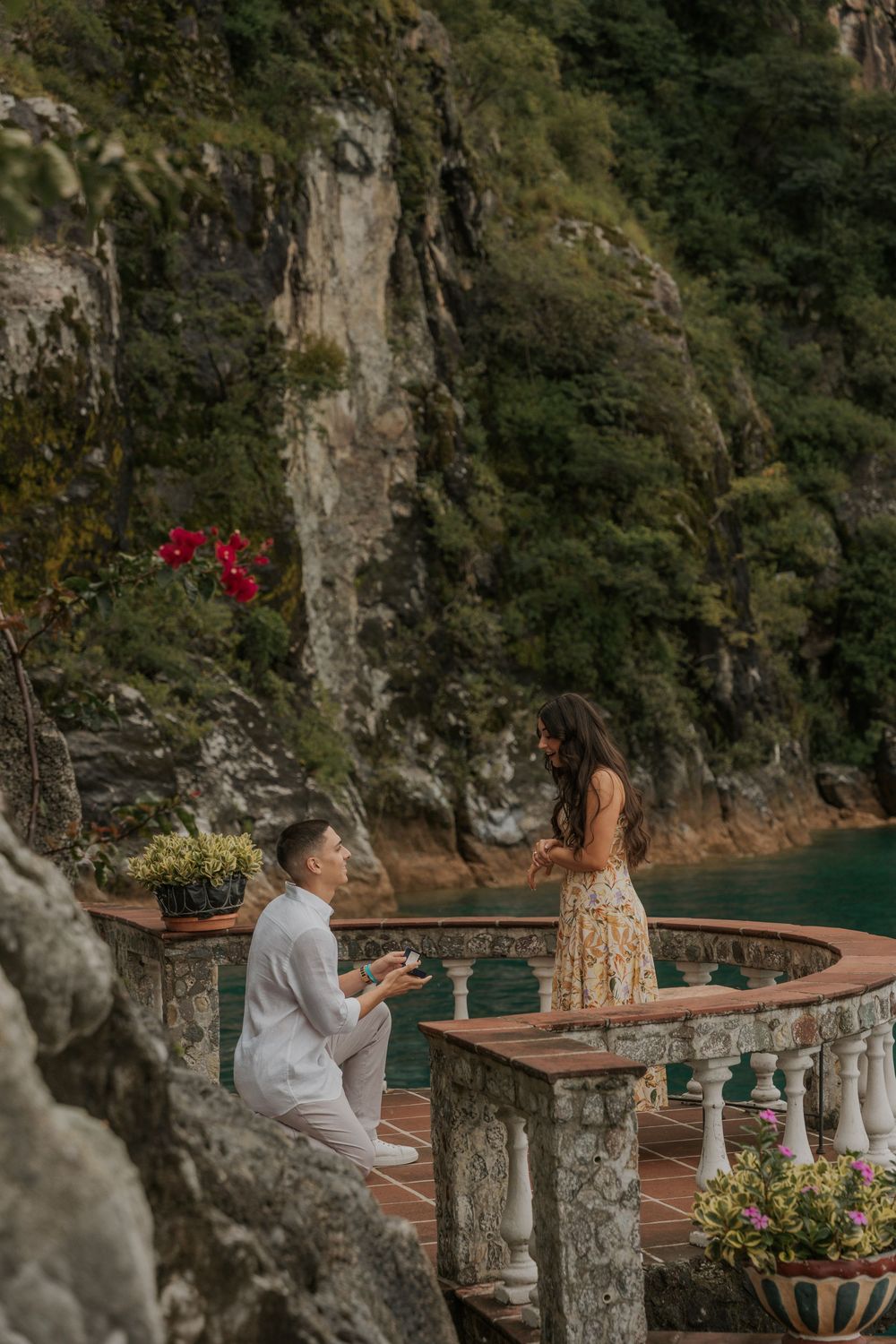 A couple shares a romantic moment on a scenic terrace overlooking the Amalfi Coast with stone columns and flowers.