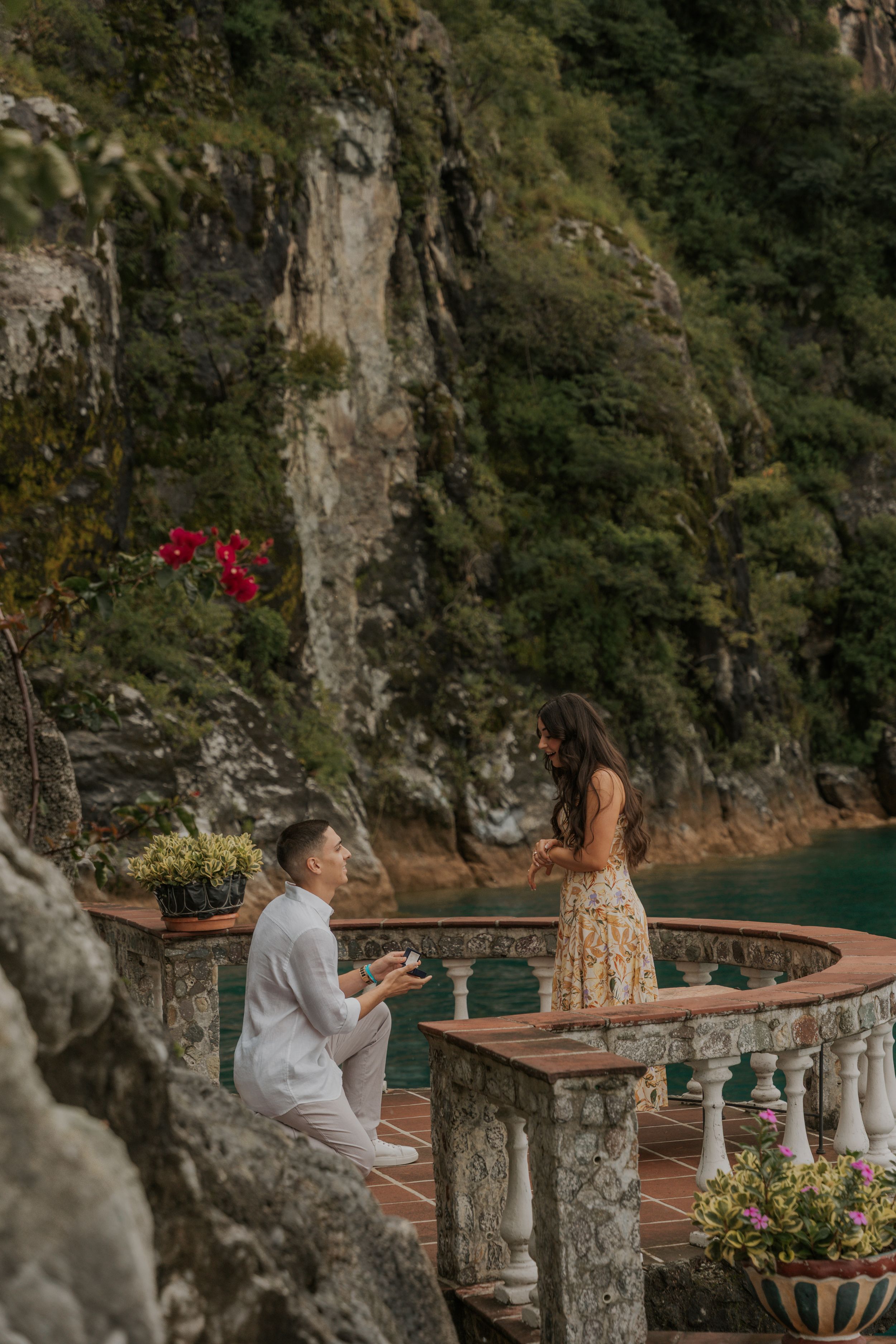 A couple shares a romantic moment on a scenic terrace overlooking the Amalfi Coast with stone columns and flowers.