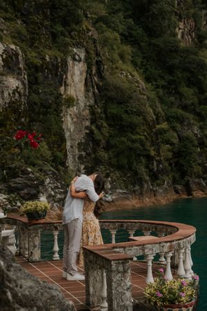 A curved stone terrace overlooks turquoise waters along a scenic coastal cliff with colorful flowers and decorative columns.