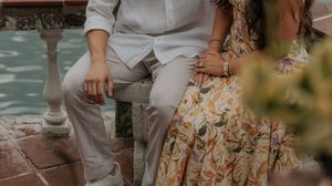 Two people sit together on a bench wearing light colored clothing and a floral dress near water.