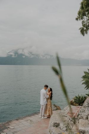 A couple shares an intimate moment on a stone terrace overlooking a misty lake with mountains in the background.