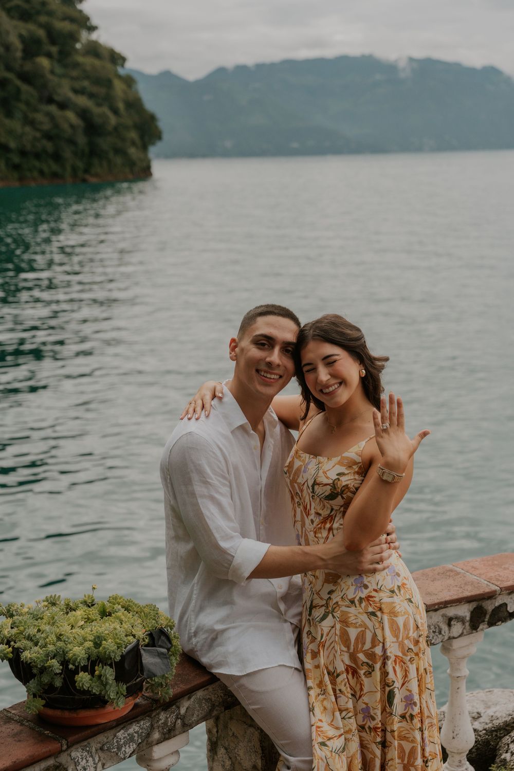 Couple embracing on a lakeside terrace with mountains and cloudy skies in the background.