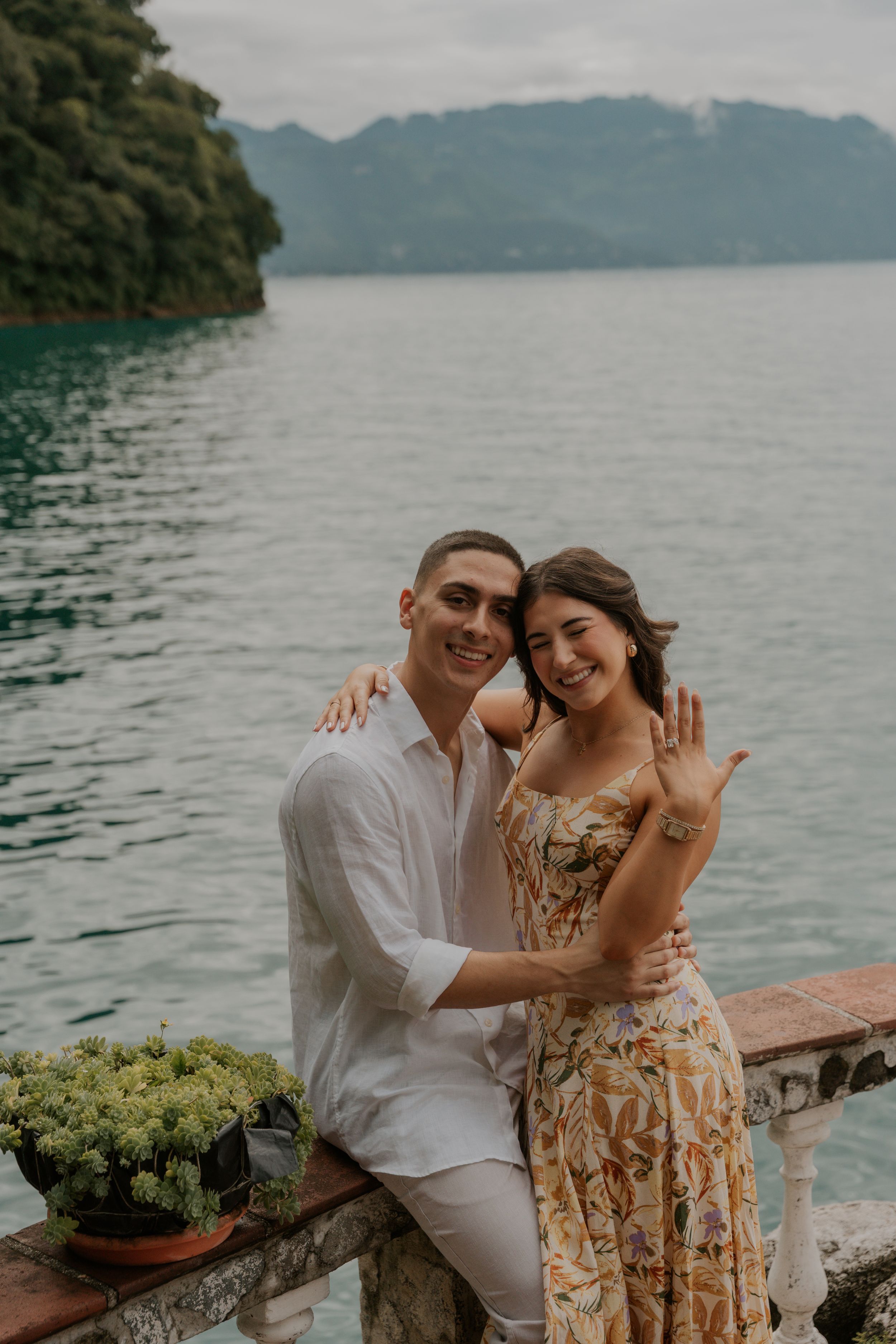Couple embracing on a lakeside terrace with mountains and cloudy skies in the background.