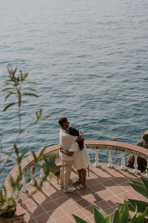 Couple embracing on a scenic terrace overlooking calm blue ocean waters at dusk.
