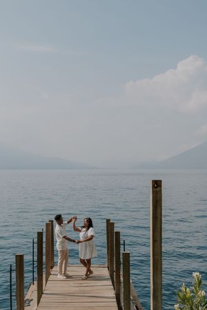 Happy couple dances on a wooden pier overlooking a serene blue lake with mountain views.
