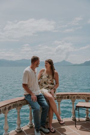 Couple sitting together on a stone balcony overlooking Lake Atitlan with mountains in the background on a cloudy day.