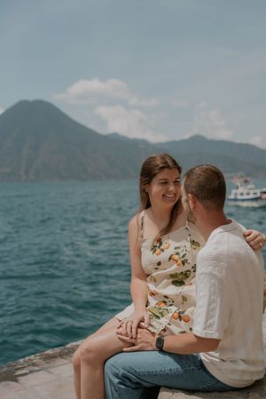 Two people sit close together on a coastal overlook with mountains and blue waters in the background.