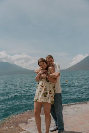 Couple embraces on a dock overlooking a scenic mountain lake with cloudy skies and turquoise waters in the background.