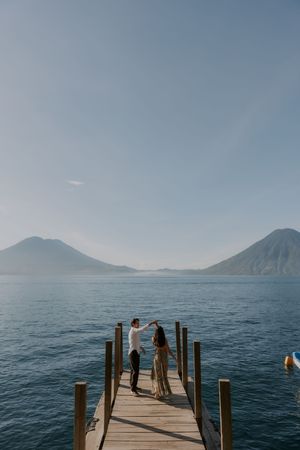 Wooden dock stretches into Lake Atitlan with distant volcanoes rising above calm blue waters in Guatemala.