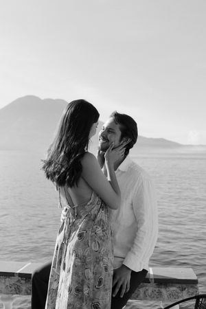 A romantic couple shares a tender kiss by the ocean with mountains in the background.