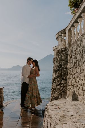 A romantic couple embraces on a stone terrace overlooking a scenic lake with mountains in the background.