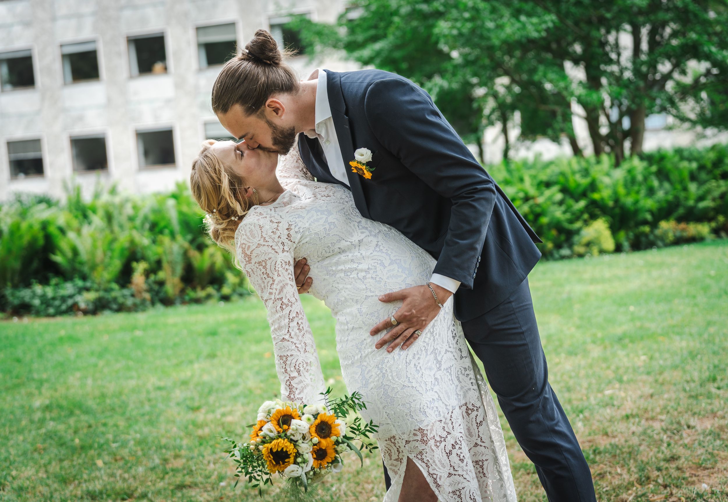 A couple in wedding attire share a romantic moment in a lush garden setting with sunflowers nearby.