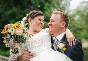 Joyful wedding couple embraces outdoors surrounded by lush greenery while holding a vibrant floral bouquet.