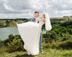 A couple in wedding attire dances together on a hilltop overlooking a scenic lake and rolling green landscape.