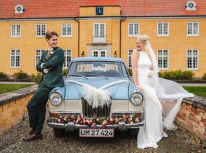 A bride and groom pose with a classic vintage car against a rustic yellow building backdrop.