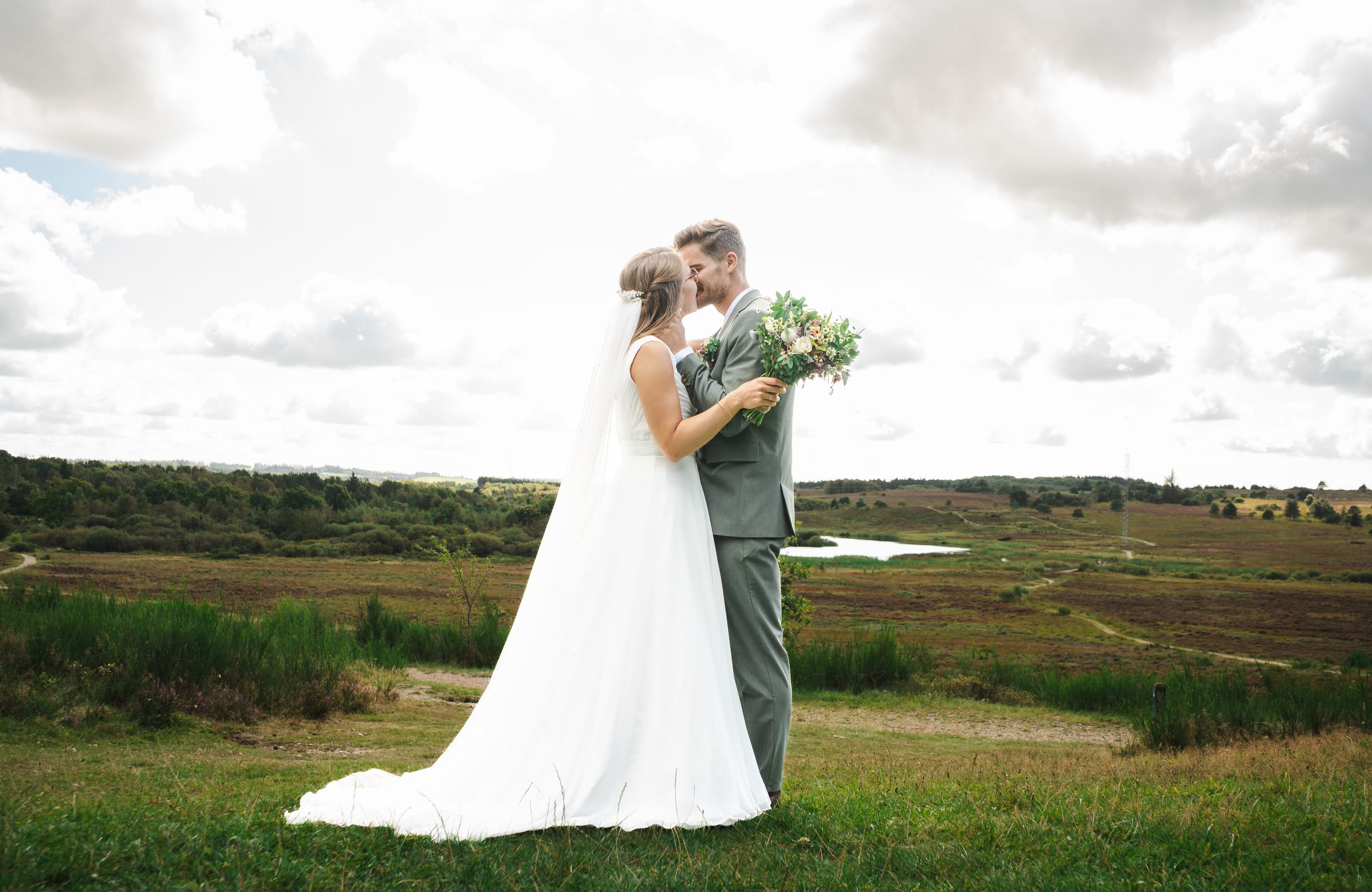 A couple in wedding attire embraces while standing in a scenic green meadow with rolling hills in the background.