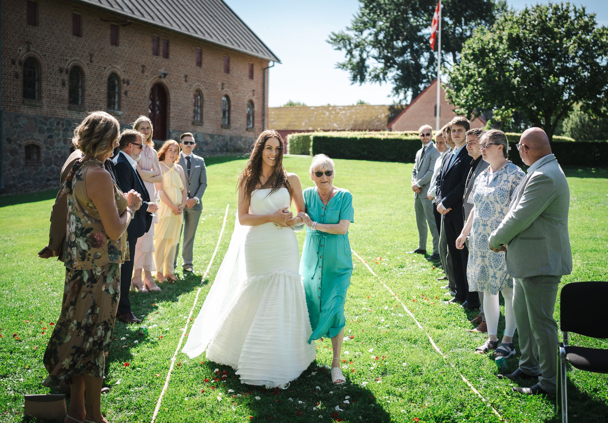 A wedding party celebrates outdoors on a sunny day with guests gathered on a green lawn near a stone building.